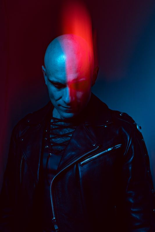 Promo portrait: a face split between red and blue light, leather jacket